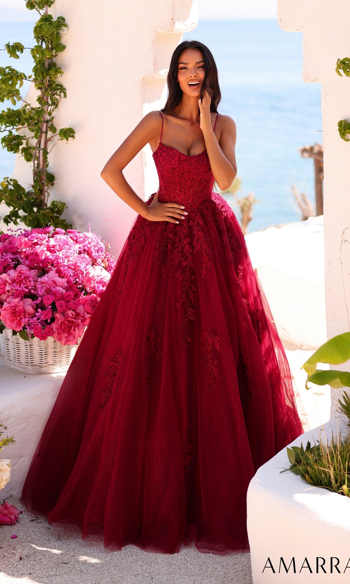 A woman poses outdoors by white walls and pink flowers with the sea in the background, wearing the Amarra 89806 long prom dress in wine.